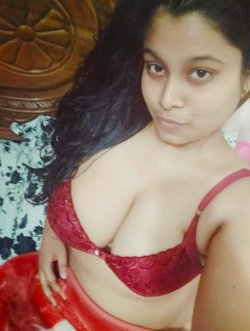 Woman with long dark hair wearing a red lace bra, posing indoors near a wooden headboard.