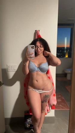 Curvy woman with curly hair and glasses takes a mirror selfie in a light blue bra and white thong, holding a phone,