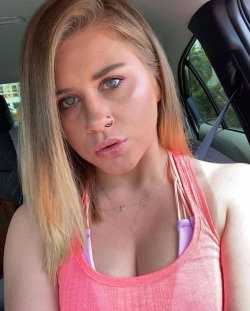 Blonde woman with nose ring in a pink tank top, taking a selfie inside a car.