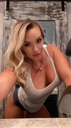 Blonde woman with curvy figure wearing a white tank top and dark bottoms, posing in front of a rustic wooden door.