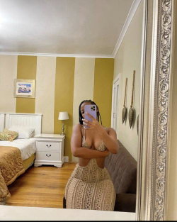 A curvy woman in a beige lace dress takes a mirror selfie in a bedroom with striped walls and wooden floors.