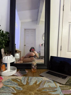 A woman with long dark hair sits on the floor in front of a full-length mirror, taking a selfie with her phone. She