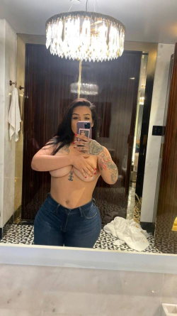 Brunette woman with tattoos takes mirror selfie in bathroom, topless with hands covering chest, wearing jeans under