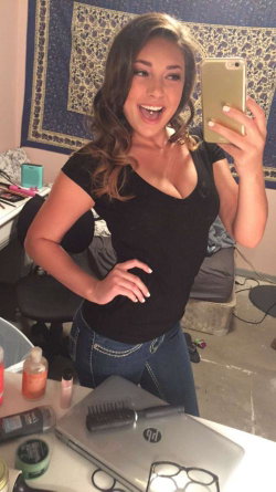 A smiling woman with wavy brown hair takes a mirror selfie in a black top and jeans, standing in a room with a desk and