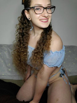 A woman with curly brown hair and glasses wears a light blue off-shoulder top and matching thong, sitting on a couch
