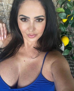 Brunette woman with defined features in a blue top, posing outdoors near lemon plants.