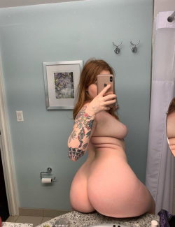 A woman with long red hair and a full sleeve tattoo takes a mirror selfie in a bathroom, showing her bare back and