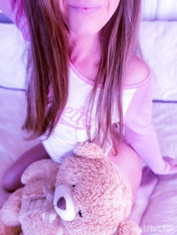 A woman with long brown hair in a white and pink off-shoulder top sits on a bed holding a teddy bear, under soft pink