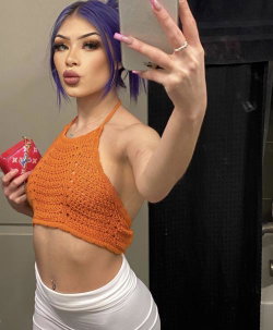 Woman with purple hair takes mirror selfie in orange crocheted crop top and white high-waisted pants, holding a red