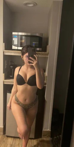 Curvy woman in black bra and patterned thong takes mirror selfie in kitchenette, holding phone with one hand.