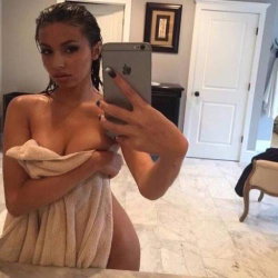 Brunette woman with curvy build takes mirror selfie holding phone, partially covered by towel in a bathroom setting.