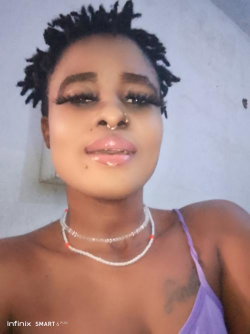 A Black woman with short dreadlocks, wearing a purple top and layered necklaces, poses in a selfie with visible chest
