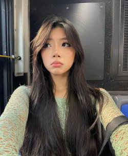 A young woman with long dark hair and a green patterned top takes a selfie inside a vehicle, looking upward with a
