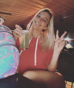 Blonde woman in a red hoodie smiles while holding a drink and making a peace sign, seated indoors with a colorful