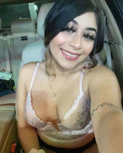 A smiling woman with dark hair and makeup sits in a car, wearing a lace bra and a necklace, taking a selfie.