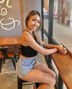 A smiling woman with long hair sits on a stool at a cafe, wearing a black crop top and denim shorts.