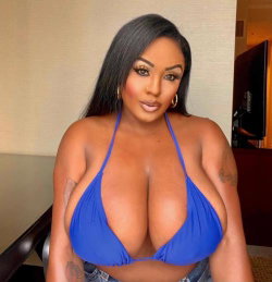 A curvy woman with long dark hair wears a blue bikini top and denim bottoms, posing indoors with visible arm tattoos.