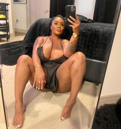 A curvy woman with dark hair takes a mirror selfie in black lace lingerie, sitting barefoot on the floor near a bed.