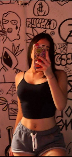 Brunette woman in black crop top and gray shorts takes mirror selfie in front of graffiti-covered wall.