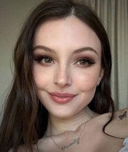 Brunette woman with long hair, subtle makeup, and a nose ring smiles softly in a close-up selfie indoors.