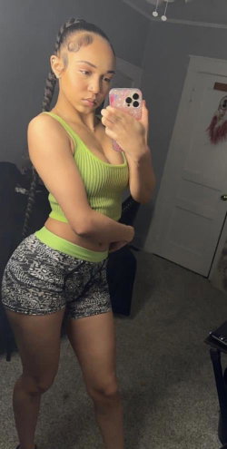 A woman with braided hair takes a mirror selfie in a lime green crop top and patterned shorts, standing in a dimly lit
