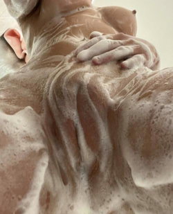 A person with bare breasts covered in soap suds, lying back with a hand on their chest in a shower setting.