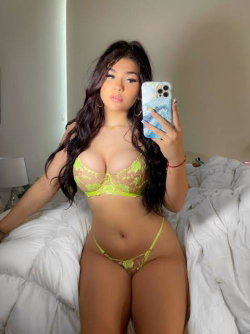A woman with long dark hair in a neon green lace bra and matching thong takes a mirror selfie on a bed.