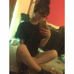 A young woman with dark hair in a bun and glasses takes a mirror selfie, wearing a black t-shirt and sitting on the