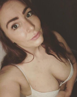 A woman with dark hair and a nose ring wears a white lace bra, posing closely in a dimly lit room.