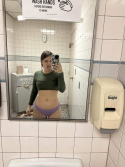 Brunette woman in a green crop top and purple thong takes a mirror selfie in a tiled bathroom, sticking out her tongue.