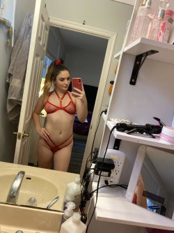 A woman with blonde hair in a ponytail takes a mirror selfie in a red lace lingerie set in a bathroom.