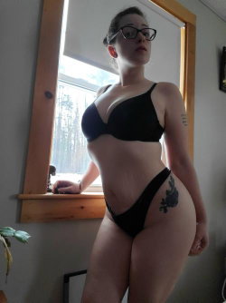 Curvy woman in black lingerie poses by a window, wearing glasses with a floral tattoo on her hip.