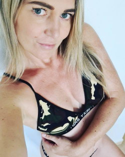 Blonde woman with blue eyes in a camouflage-patterned bra, posing closely to the camera against a plain white