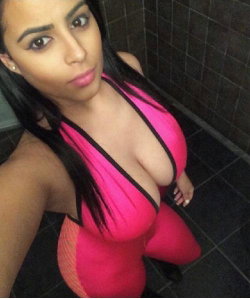 Brunette woman in a bright pink and black bodysuit poses in a tiled shower, taking a selfie with visible arm and phone