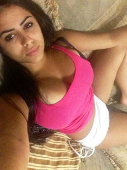 Brunette woman with curvy build lying on a couch, wearing a pink crop top and white shorts, taking a selfie.
