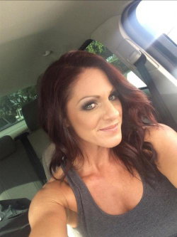 Woman with dark wavy hair and makeup takes a selfie in a car, wearing a gray tank top.