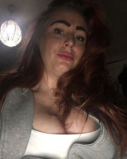 A woman with long red hair and a nose ring wears a white top under a gray zip-up hoodie, taking a close-up selfie