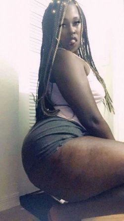 Curvy Black woman with long braids wearing a white tank top and dark shorts, sitting on a surface indoors.