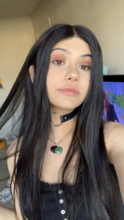 A young woman with long dark hair, wearing a black choker and a buttoned top, takes a selfie indoors.