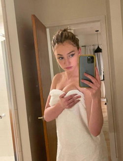 A young woman with light brown hair in a messy bun takes a mirror selfie, wrapped in a white towel, holding her phone