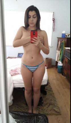 Brunette woman with curvy build takes mirror selfie in striped thong, standing barefoot in bedroom.