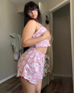 Curvy woman in pink patterned crop top and shorts, wearing glasses, posing indoors with arm across chest.
