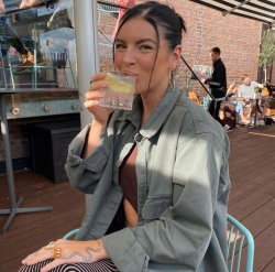 A woman with dark hair tied back sits outdoors, sipping a drink from a glass while wearing a loose olive jacket over a