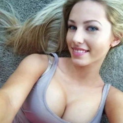 Blonde woman with blue eyes lying on carpet, wearing a gray tank top, smiling at the camera in a close-up selfie.