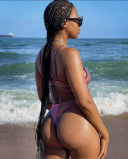 Curvy woman with long braids and sunglasses in a purple bikini, standing on a beach facing the ocean.