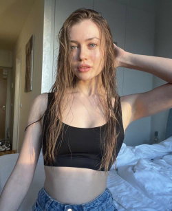 A slim woman with wet hair wears a black crop top and jeans, posing in a bedroom with one hand in her hair.