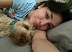 A woman with dark hair lies on a couch, smiling softly while cuddling a small fluffy dog.