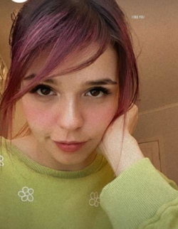 A young woman with purple-tinted hair and brown eyes wears a green sweater with floral details, resting her head on her