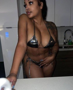 Curvy woman in metallic bikini with visible tattoos poses in a kitchen, hand resting on counter.