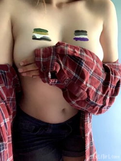 A woman with bare breasts covered by painted shapes, wearing an open plaid shirt and dark shorts, posing against a blue